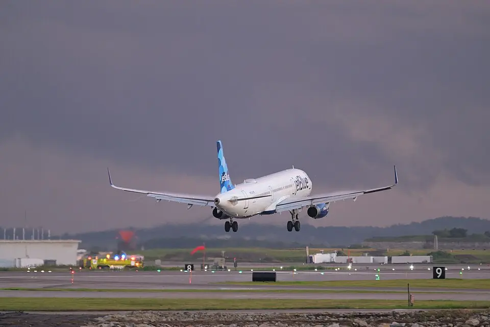 JetBlue Airways Airbus A321 climbing after takeoff at dusk with emergency vehicles staged nearby
